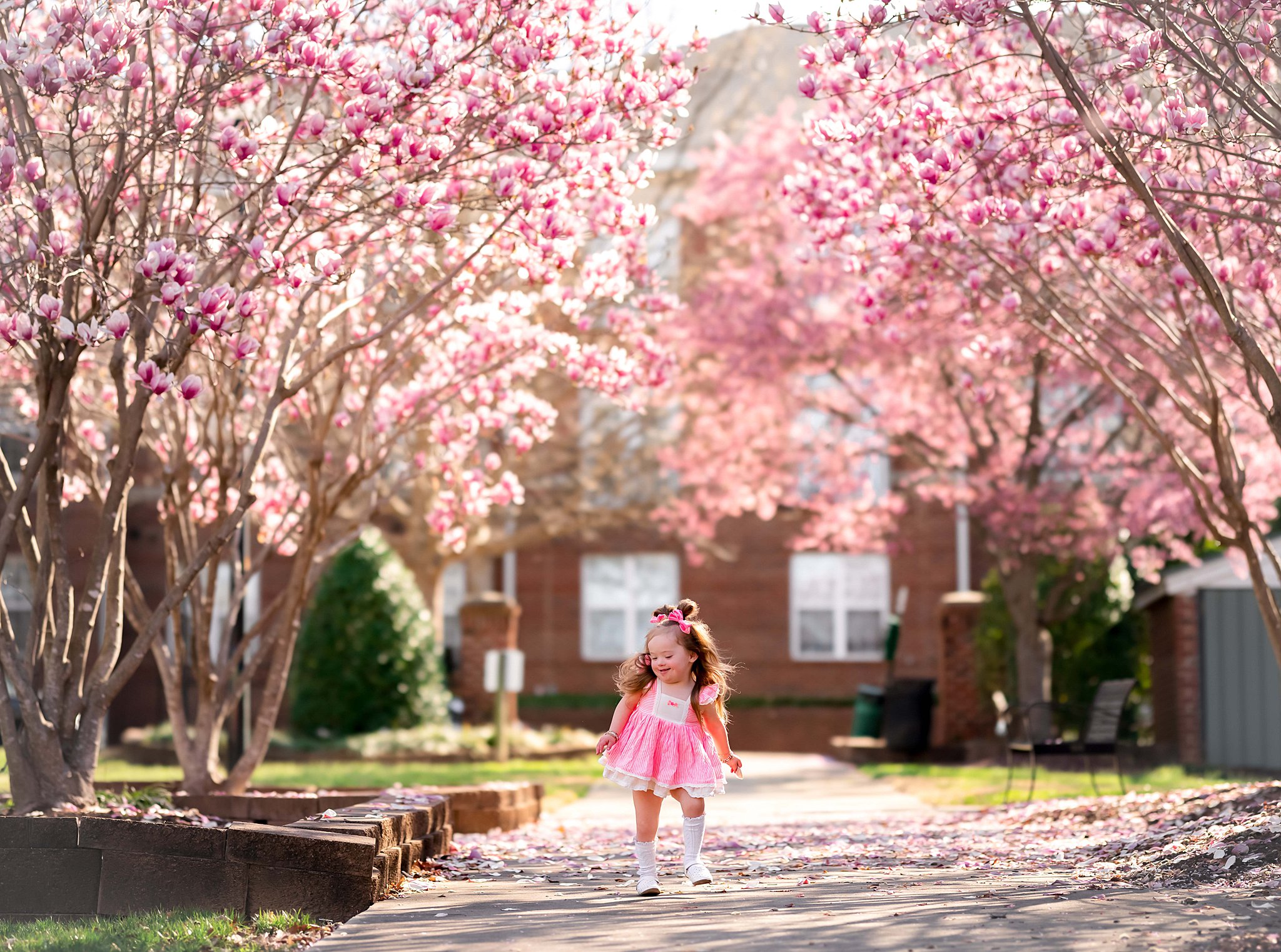 A young girl in a classic pink dress plays under the blooming pink flowers of trees Children’s Boutique Raleigh NC