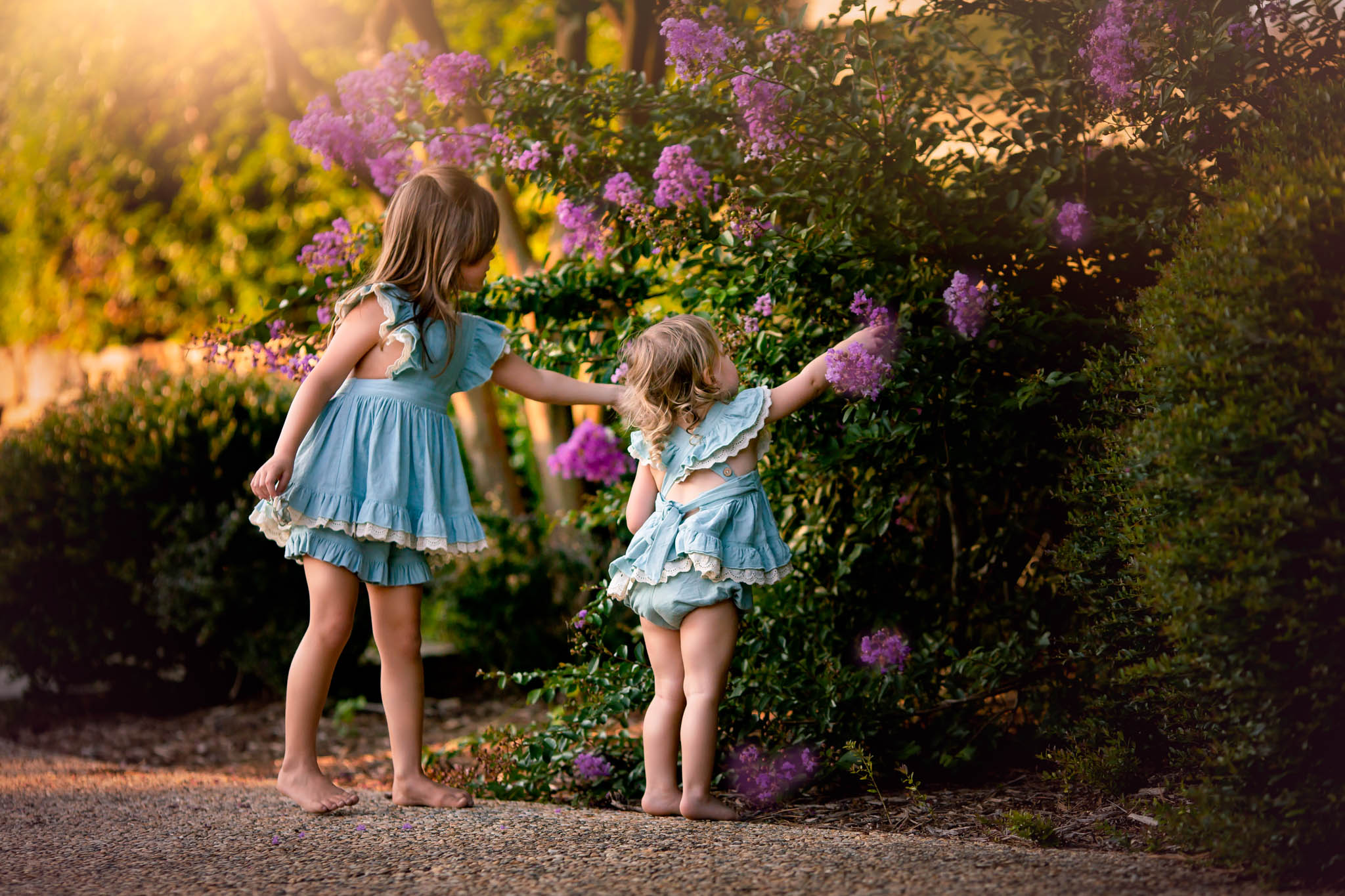 Two sisters in matching blue dresses pick purple flowers barefoot in a park pattywhacks