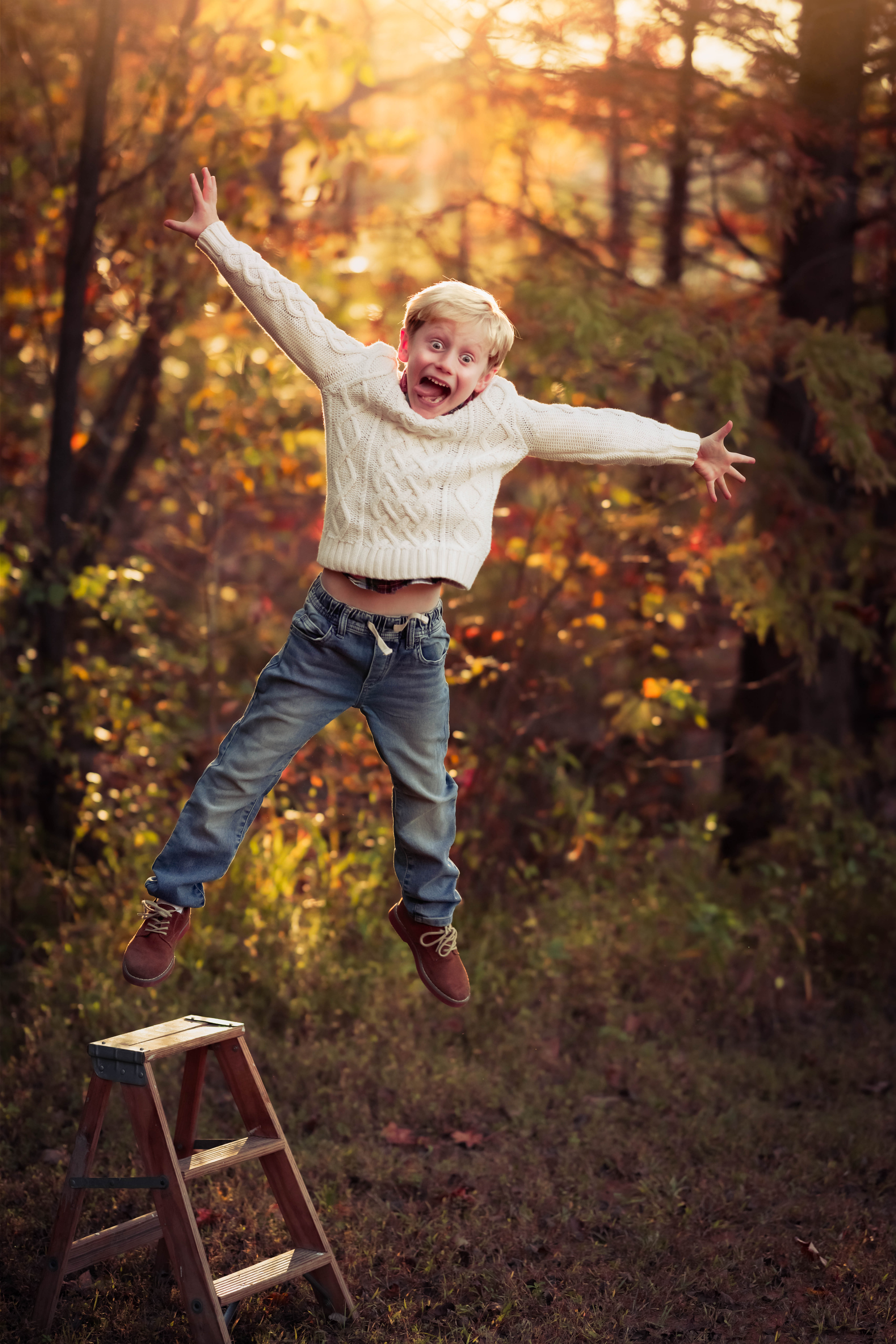A young boy jumps off a wooden step ladder with arms out making a funny face in a forest raleigh toy stores