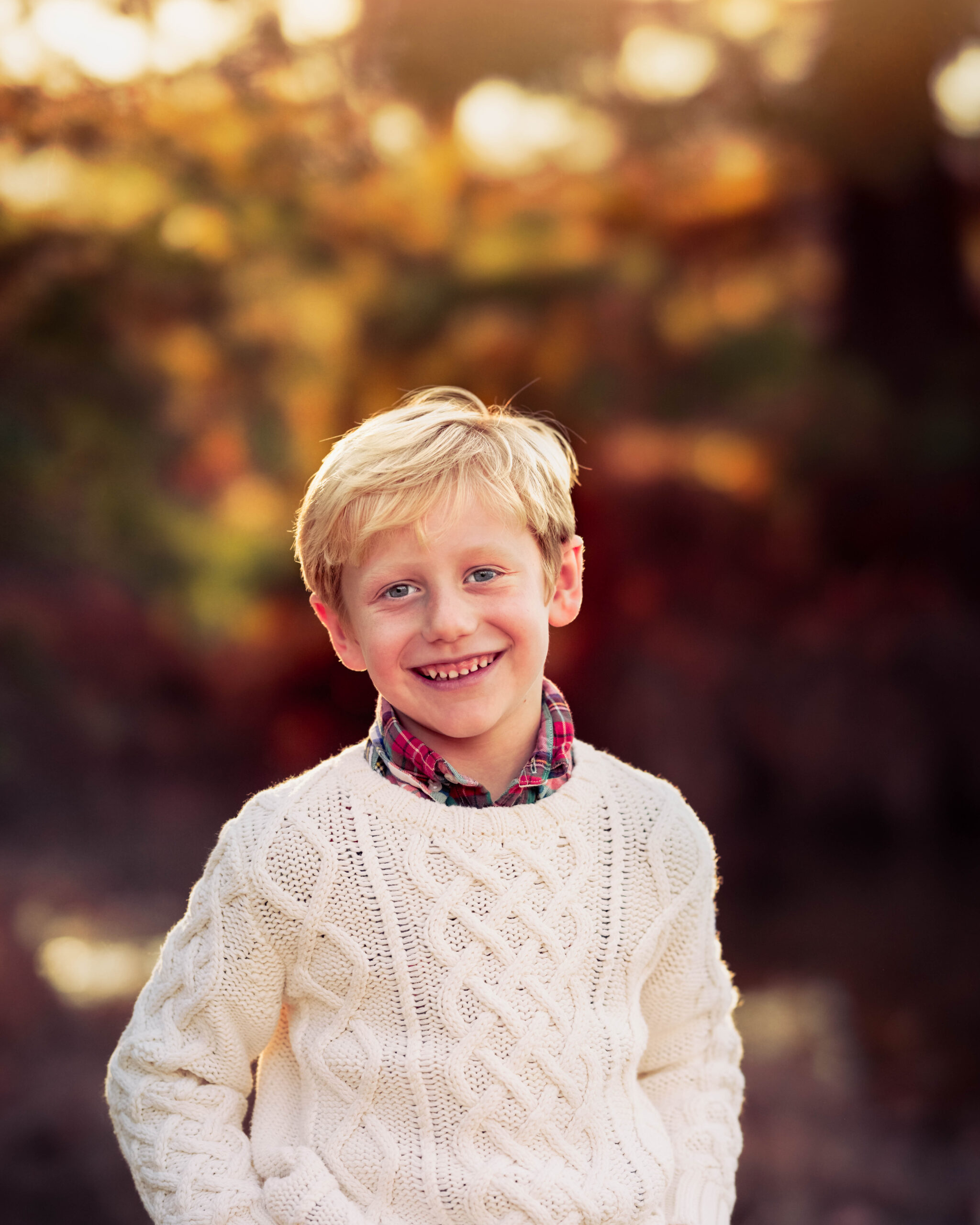 A young boy in a white knit sweater stands in a park at sunset