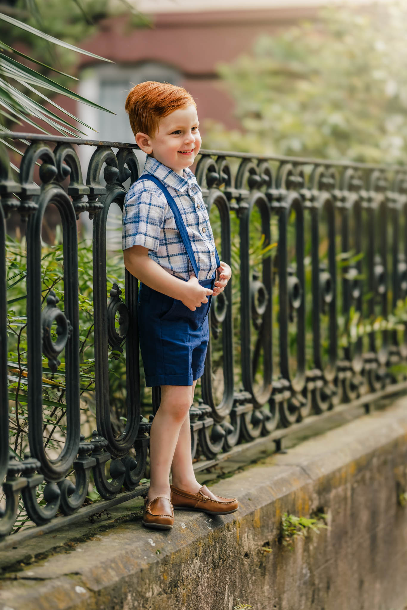A young boy stands on a fence holding his blue suspenders and leaning on an iron fence things to do in asheville with kids