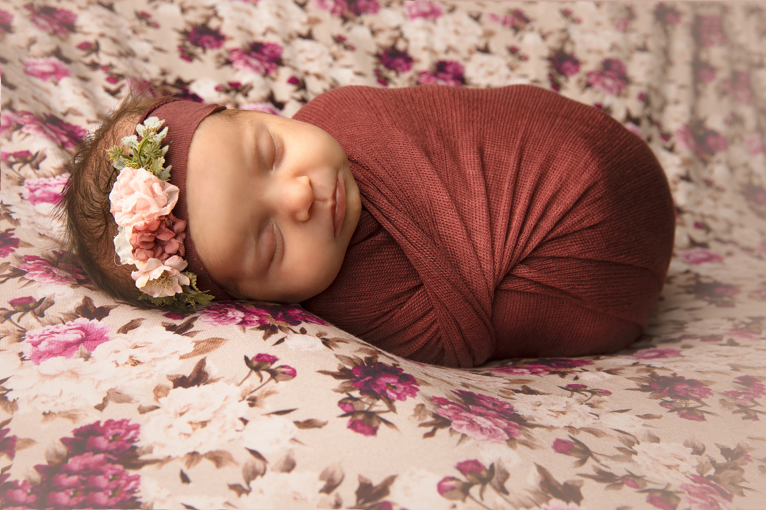A newborn baby sleeps in a red swaddle on a floral pattern bed with a flower headband