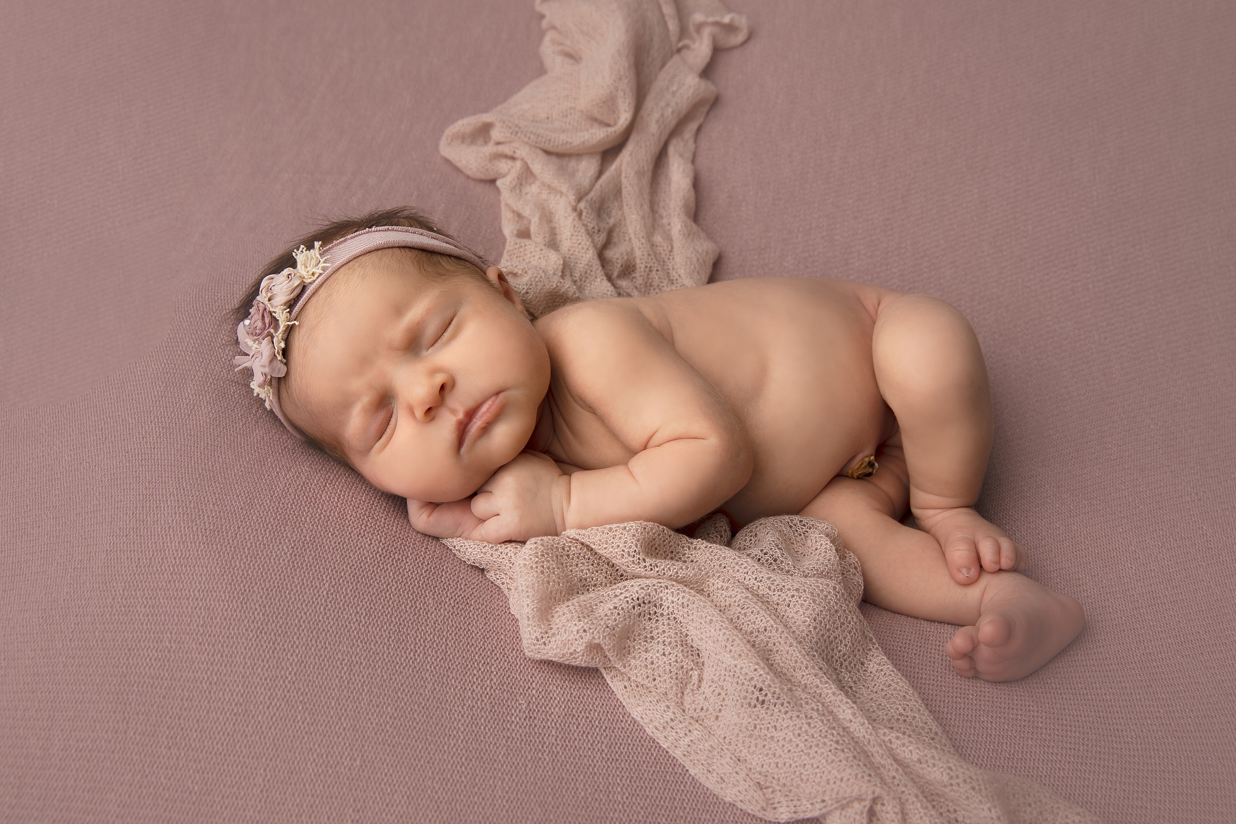 A newborn baby sleeps on its side on a pink bed wearing a headband raleigh nannies