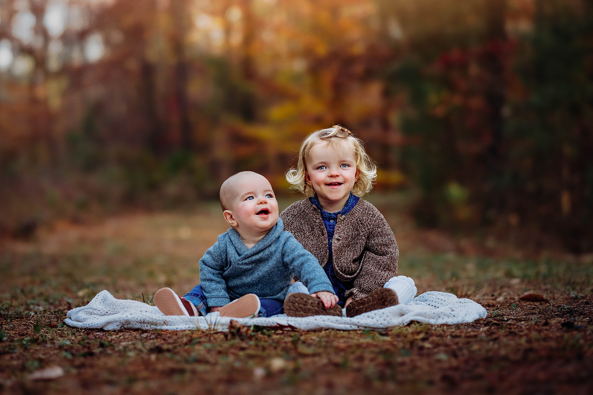 Two sibling sit together on a blanket in a park in sweaters