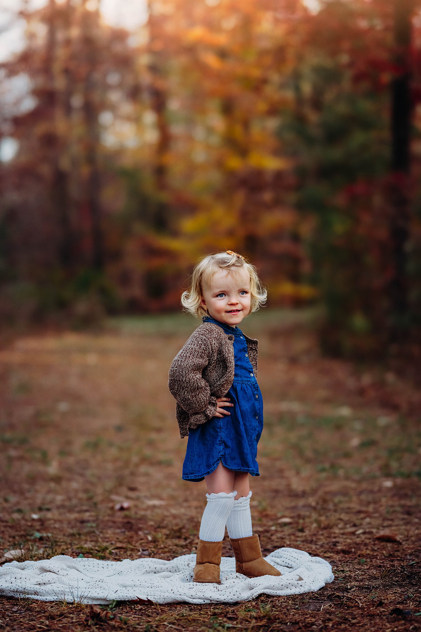 A young girl stands with hands on her hips in a denim dress in a park in fall raleigh pediatric dentists