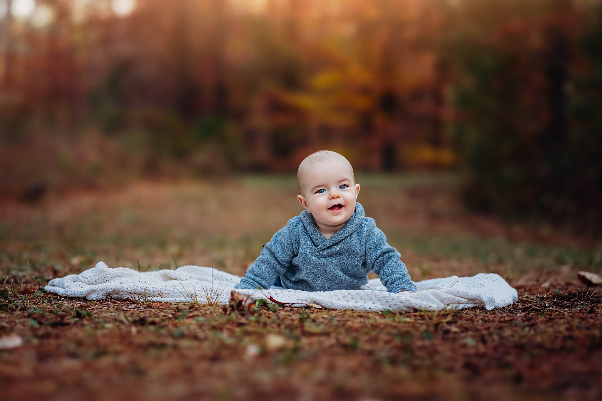 A toddler boy lays on a picnic blanket in a park in fall wearing a grey sweater raleigh pediatric dentists