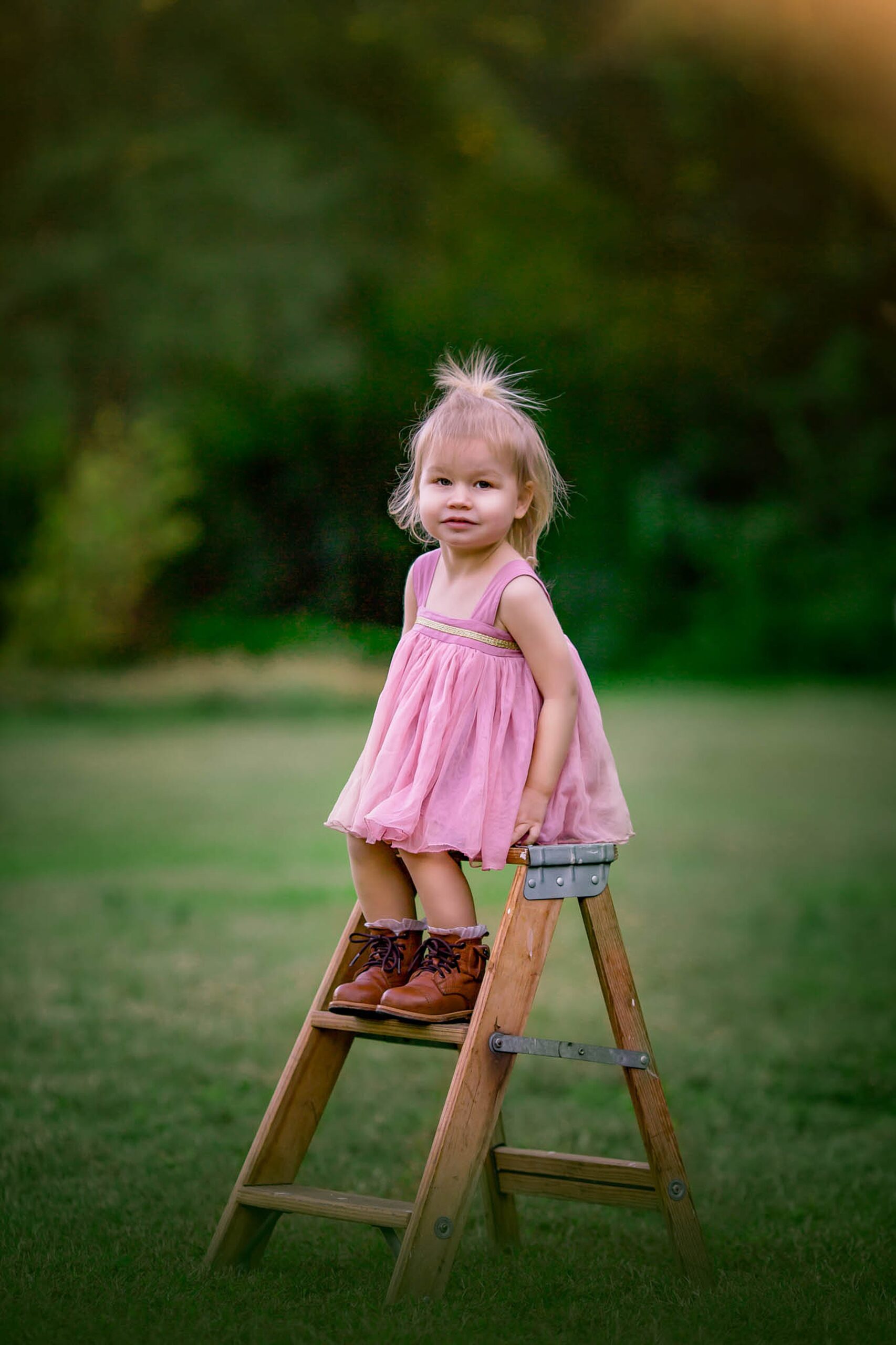 A toddler girl in a pink dress and boots sits on a wooden step stool in a park raleigh playgrounds