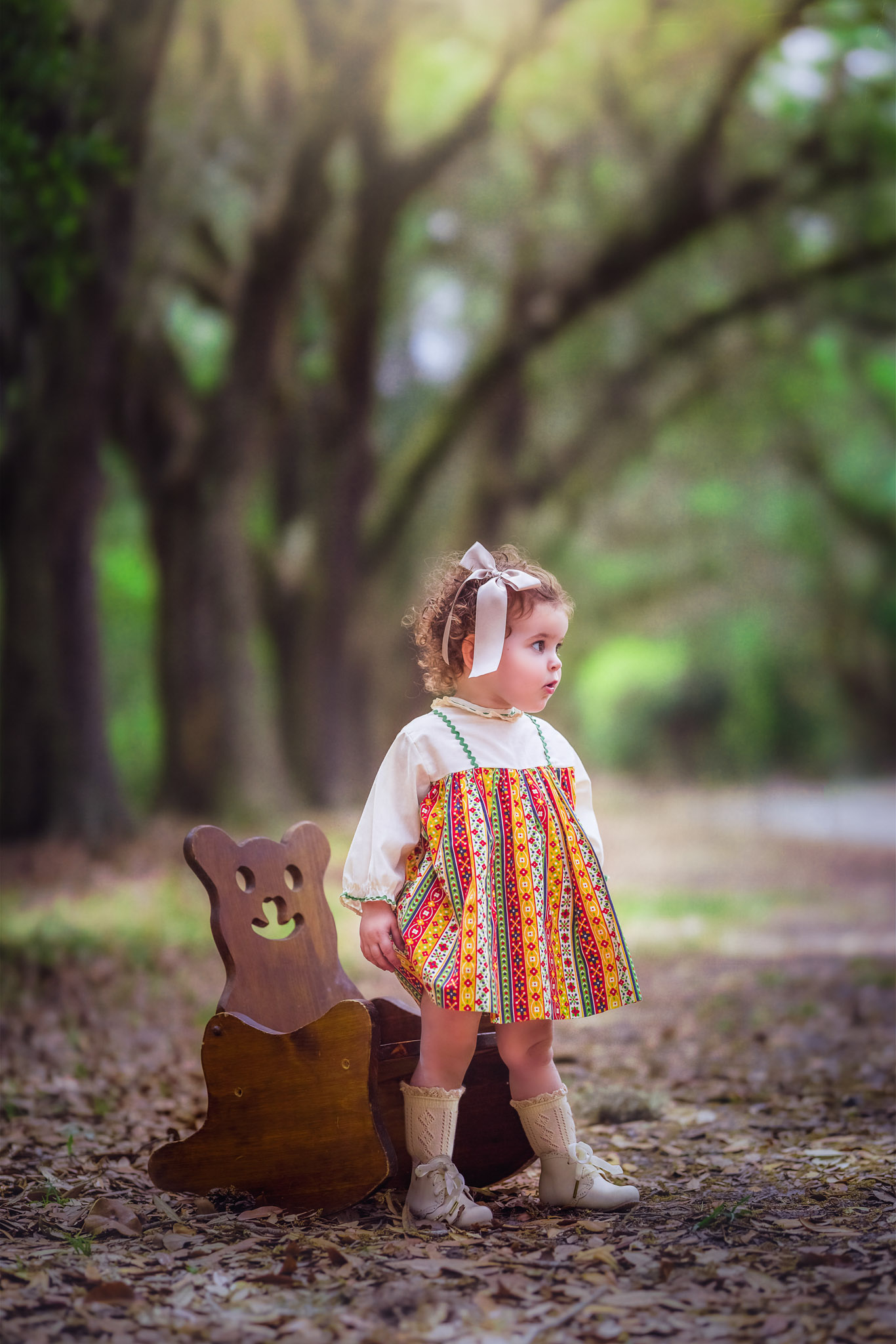 A young girl in a colorful patterned dress and high socks stands in a park in front of a wooden teddy bear chair raleigh preschools
