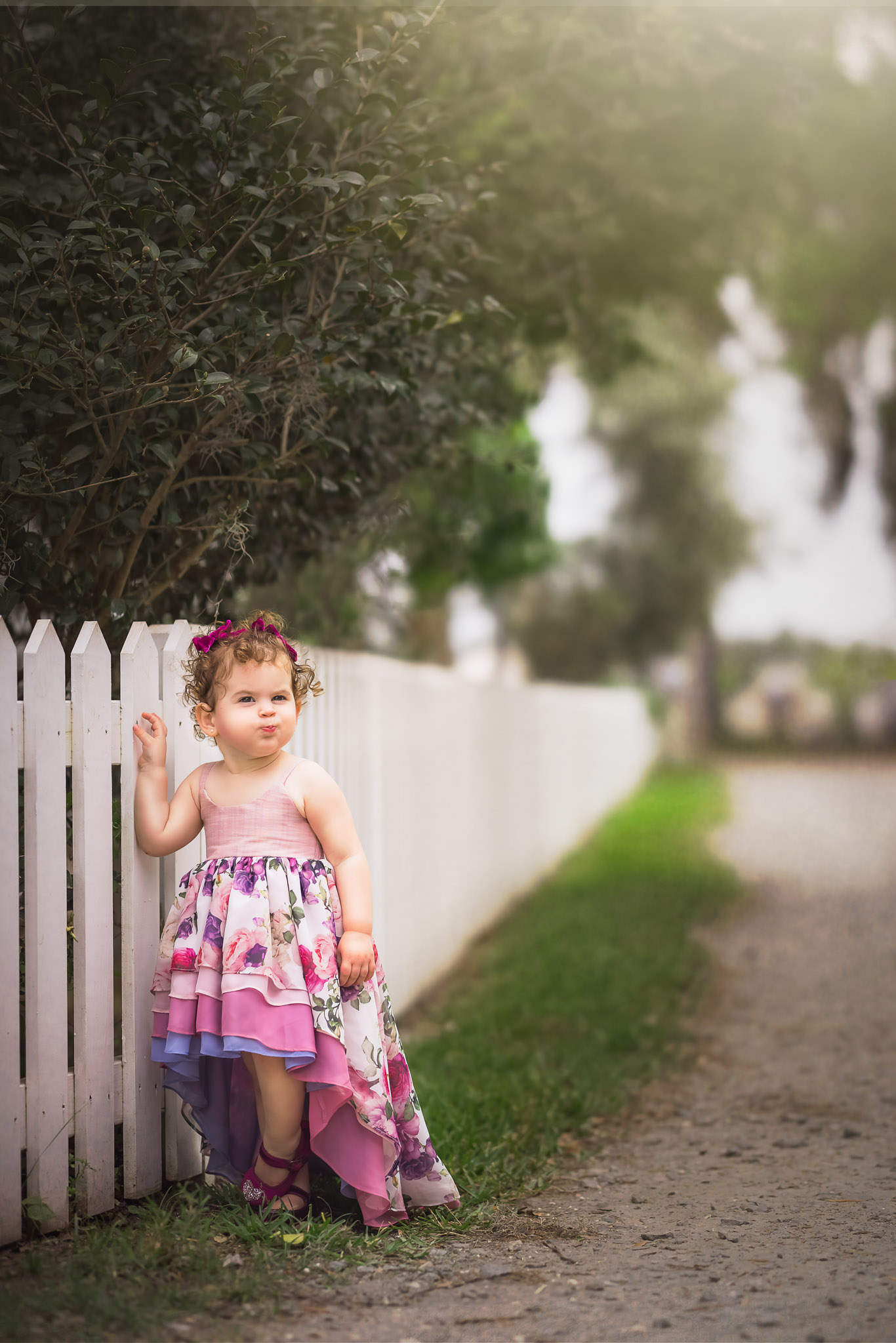 A young toddler girl in a colorful pink dress stands with a hand on a white picket fence raleigh preschools