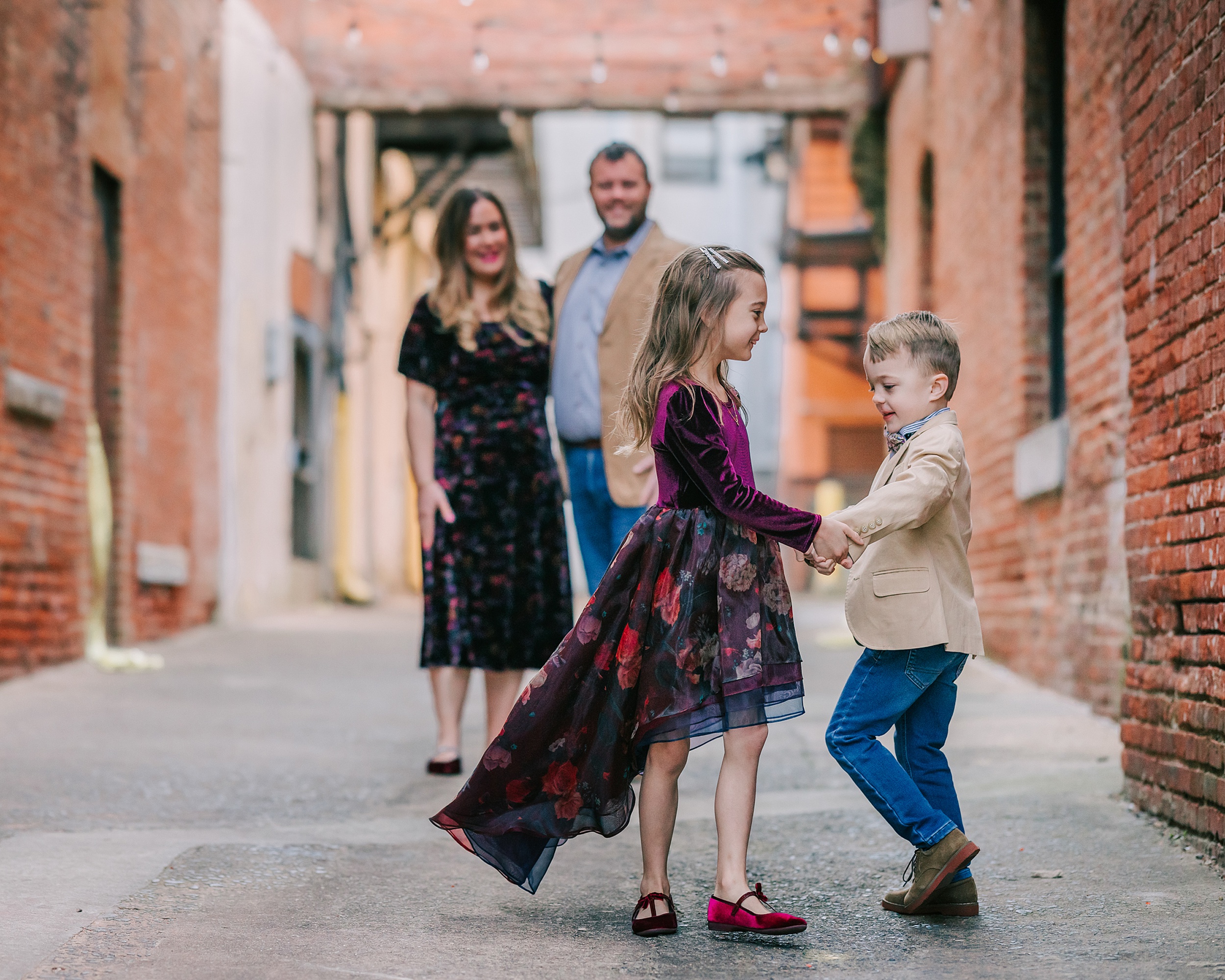 A young brother and sister dance in a alley while mom and dad in matching outfits stand behind them