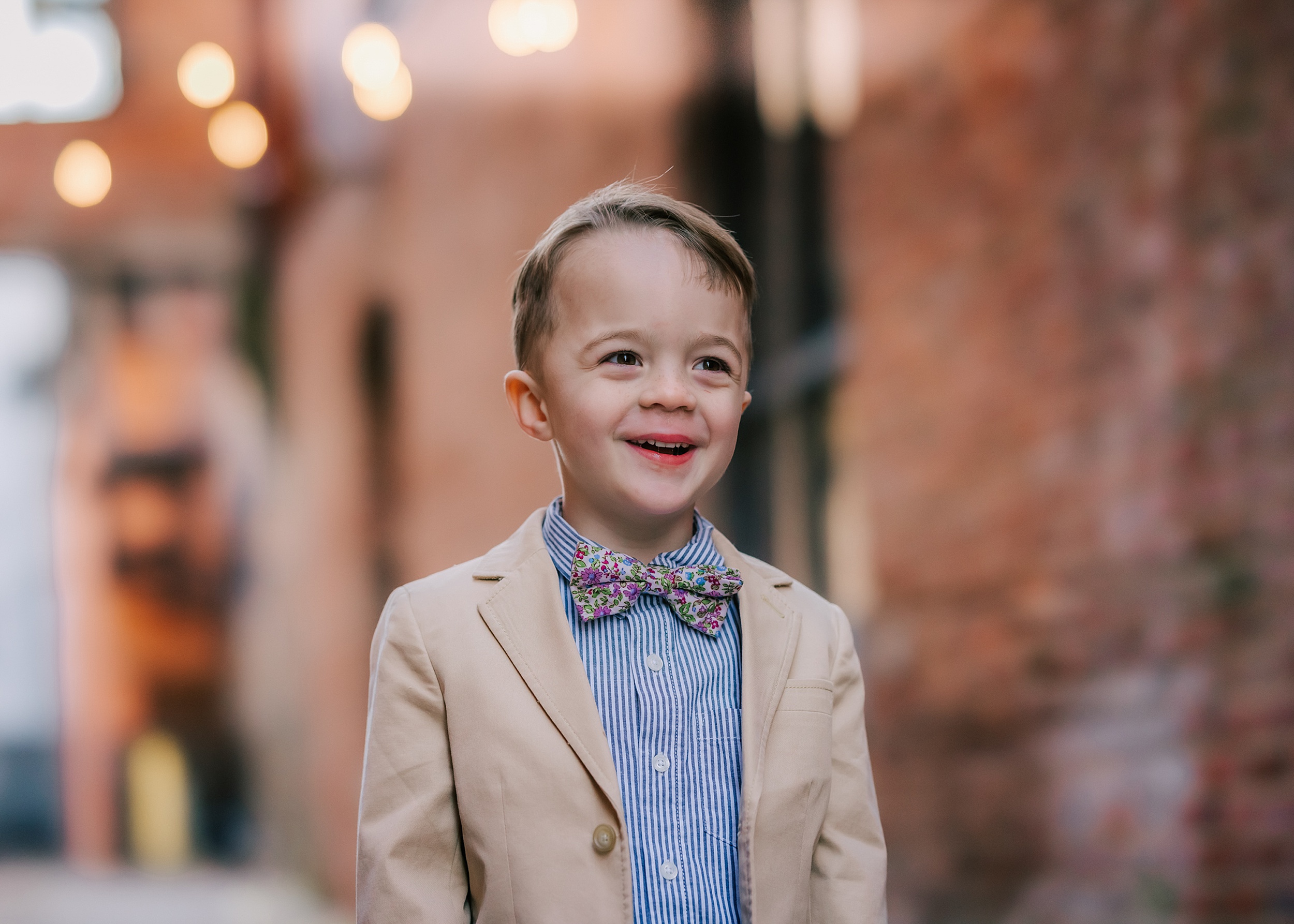 A young boy stands in an alley in a tan suit jacket, blue stripe shirt and bowtie raleigh youth sports